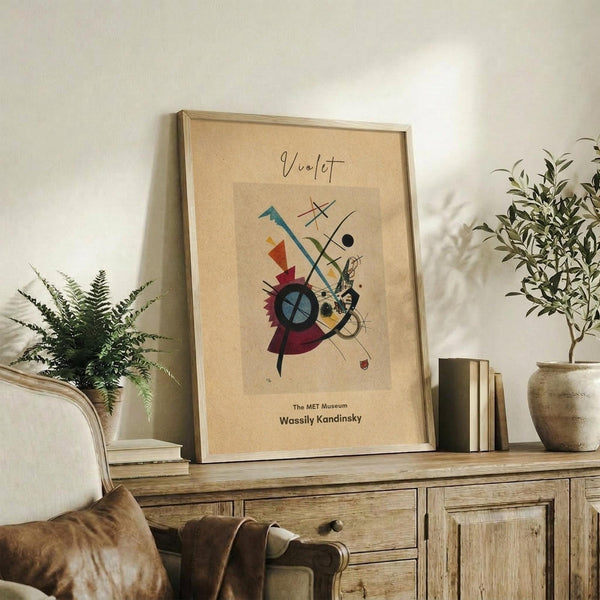 May include: Framed abstract art print in warm beige tones featuring geometric shapes, lines, and a circular motif in blues, reds, and yellows. Print sits on rustic wooden sideboard alongside potted plants, books, and ceramic vase. Cream upholstered chair with brown throw pillow visible in foreground. Natural lighting creates soft shadows on white wall background.