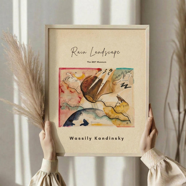 May include: Hands holding a framed abstract art print featuring flowing organic shapes in watercolor tones of red, yellow, blue, and brown on cream background. Light wood frame with cream matting. Person wearing beige ruffled sleeve top stands by window with dried pampas grass visible to the left.