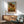 May include: Framed sunflower artwork hanging on white wall above wooden credenza with table lamp. Print shows three large sunflowers with yellow petals and dark centers against blue sky and golden field. Modern living room setting with mint green upholstered chair, round wooden coffee table, and natural sunlight casting shadows across the scene.