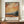 May include: Framed poster featuring a Van Gogh-inspired landscape painting with swirling orange and yellow sky, rolling hills, and dark cypress trees beside a small building. The artwork has visible brushstroke texture and warm earth tones. Below the painting is text in French about contemporary art inspired by a master artist, along with Van Gogh's name and an English quote about dreaming and painting. The frame sits on a wooden sideboard next to a white ceramic vase and books in a cream-walle