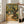 May include: A framed Van Gogh-style poster showing an olive tree with thick, gnarled trunk and golden-green foliage painted in heavy impasto brushstrokes. A small house sits in the background along a winding path. The artwork features warm yellows, greens, and earth tones in the characteristic swirling style. Someone holds the light wood-framed print in a bright living room with white walls, large windows, and a beige sofa.
