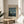 May include: Framed poster featuring Van Gogh's The Starry Night painting with swirling blue sky, yellow stars and moon, dark cypress tree, and village below. Museum-style exhibition poster with "VAN GOGH" text and "1889" date in thin wooden frame, displayed on round wooden side table next to beige upholstered chair. Soft natural lighting creates shadows on white wall, with green plant leaves visible in corner.