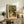 May include: Framed Van Gogh olive trees print with swirling brushstrokes in golden yellows and greens, showing a gnarled tree beside a small cottage on a cobblestone path. The artwork sits on a weathered wood sideboard next to a fern plant, table lamp, ceramic vase, and stacked books in a warm, neutral living room setting.