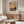 May include: Van Gogh-inspired landscape poster with swirling orange and yellow sky, dark cypress trees, and rolling golden fields, displayed in thin wooden frame above walnut credenza. Modern living room setting features cream curved chair, glass oval coffee table, brass floor lamp, and white table lamp on console.