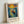 May include: Framed art print in light wood frame leaning against white wall. The artwork shows a figure in blue clothing standing in a wooden rowboat on water, with swirling yellow sun and colorful sky in Van Gogh's distinctive post-impressionist style. Blue mountains and reflections visible on water surface. Text below reads "Vincent Van Gogh" with French description and quote about dreaming and painting.