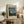 May include: Framed Van Gogh Starry Night museum poster print displayed on rustic wooden sideboard alongside fern plant, ceramic vase, and stacked books. Poster features swirling blue night sky with yellow stars and moon over village silhouette, housed in light wood frame. Styled in warm, neutral living room setting with beige armchair and cream walls.