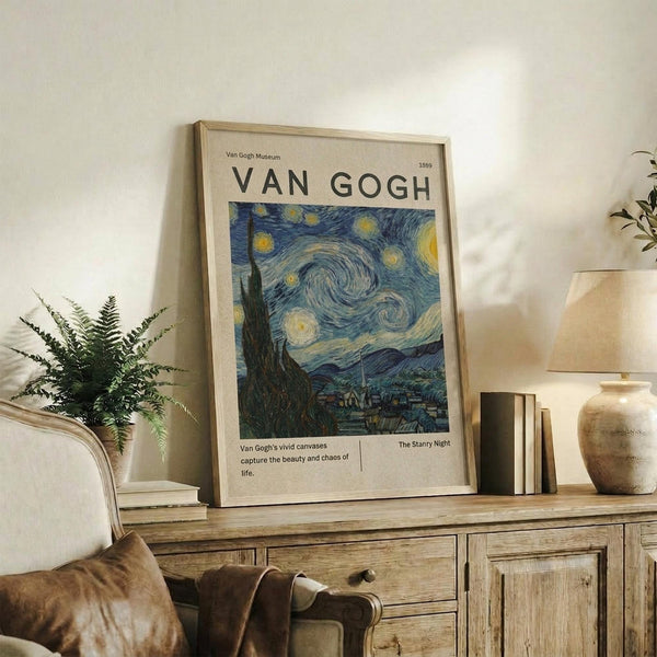 May include: Framed Van Gogh Starry Night museum poster print displayed on rustic wooden sideboard alongside fern plant, ceramic vase, and stacked books. Poster features swirling blue night sky with yellow stars and moon over village silhouette, housed in light wood frame. Styled in warm, neutral living room setting with beige armchair and cream walls.