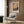 May include: A framed Van Gogh-inspired landscape poster featuring cypress trees against an orange sunset sky with rolling hills, displayed above a beige upholstered armchair with geometric patterned pillow. The artwork is mounted on wood-paneled walls in a modern living space with brass floor lamp, dark side table with open book, and white curtains.
