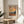 May include: Framed Van Gogh-inspired landscape poster with swirling orange and blue sky, cypress trees, rolling golden hills, and distant mountains displayed on a round wooden side table next to a beige upholstered chair. Natural sunlight casts geometric shadows across white walls. Green plant leaves visible in corner.