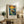 May include: Framed Van Gogh-inspired poster featuring a figure in a wooden boat on swirling blue water under a dramatic yellow sun and colorful sky, displayed on a rustic wooden sideboard alongside a fern plant, books, ceramic vase, and lamp in a cozy living room setting with neutral tones and natural lighting.