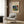 May include: Beige armchair with geometric pillow in modern living room setting. Framed art poster on wood-paneled wall features colorful abstract painting with green, blue, red, and yellow brushstrokes in expressionist style. Gold floor lamp and dark round side table with open book complete the minimalist interior scene.
