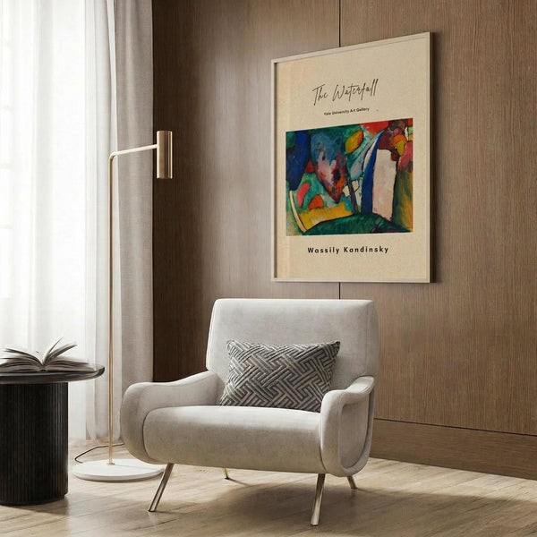 May include: Beige armchair with geometric pillow in modern living room setting. Framed art poster on wood-paneled wall features colorful abstract painting with green, blue, red, and yellow brushstrokes in expressionist style. Gold floor lamp and dark round side table with open book complete the minimalist interior scene.