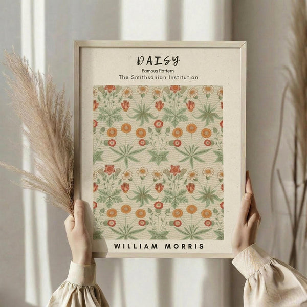 May include: Woman in cream clothing holding framed botanical print featuring William Morris daisy pattern in orange, green, and red florals. Light wooden frame, dried wheat stems visible, soft natural lighting against neutral curtained background.