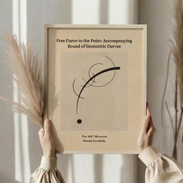 May include: Person in cream sweater holding framed abstract art print featuring curved black lines and geometric shapes on beige background. Natural lighting, dried pampas grass visible, neutral tones throughout scene. Wooden frame, minimalist home decor setting.