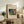 May include: Framed art print featuring an abstract waterfall painting with vibrant blues, greens, reds, and yellows in an expressionist style, displayed on a rustic wooden console table alongside books, a ceramic vase, and potted fern in a neutral-toned living room setting with cream walls and beige upholstered furniture.