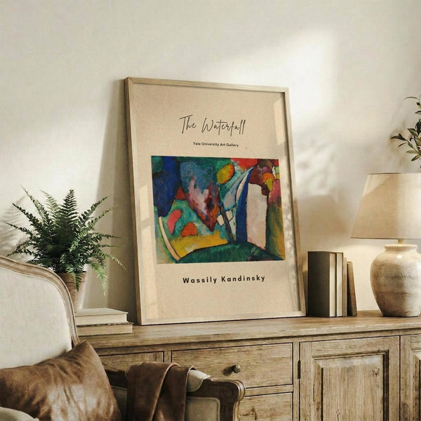 May include: Framed art print featuring an abstract waterfall painting with vibrant blues, greens, reds, and yellows in an expressionist style, displayed on a rustic wooden console table alongside books, a ceramic vase, and potted fern in a neutral-toned living room setting with cream walls and beige upholstered furniture.
