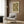 May include: Framed botanical poster with green and cream chrysanthemum floral pattern displayed on wood-paneled wall above modern beige armchair with geometric pillow. Brass floor lamp and dark side table with open book visible. Warm, contemporary interior setting with natural lighting from window.