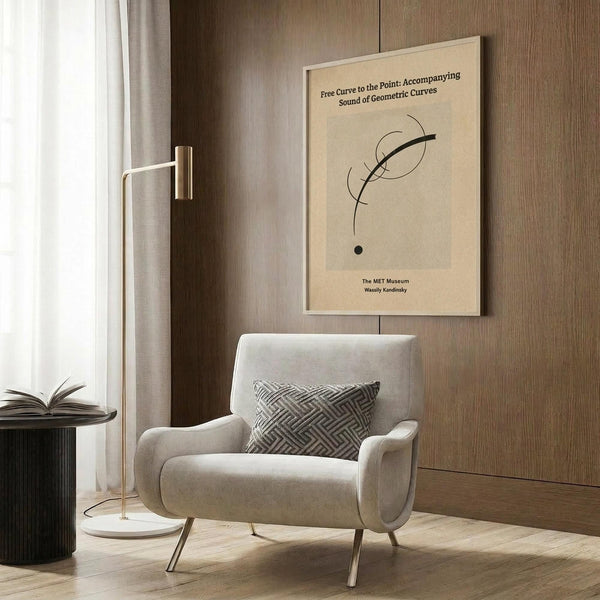 May include: Abstract minimalist art print displayed on wood paneled wall above cream upholstered armchair. Poster features curved black lines and small black dot on beige background with text about geometric curves and sound. Modern living room setting with geometric patterned pillow, brass floor lamp, and dark round side table with open book.