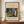 May include: Framed abstract art poster with vibrant blues, greens, oranges and reds in Kandinsky's distinctive style, displayed on wooden sideboard next to white ceramic vase and books. Hands hold large framed print against white paneled wall in elegant room setting.