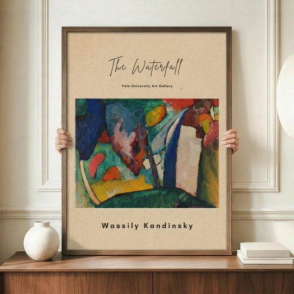 May include: Framed abstract art poster with vibrant blues, greens, oranges and reds in Kandinsky's distinctive style, displayed on wooden sideboard next to white ceramic vase and books. Hands hold large framed print against white paneled wall in elegant room setting.