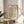May include: Framed botanical art print featuring William Morris daisy pattern with green leaves and orange-red flowers on cream background, displayed on light wood round side table next to beige upholstered chair, with plant leaves visible and natural sunlight streaming across white walls.