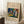 May include: A framed art print leaning against a white wall on a textured white rug. The poster features a colorful abstract painting with vibrant blues, greens, reds, yellows and white brushstrokes on a beige background with black text above and below the artwork. A white coffee cup sits on a wooden block nearby, and a beige knitted blanket is partially visible in the corner.