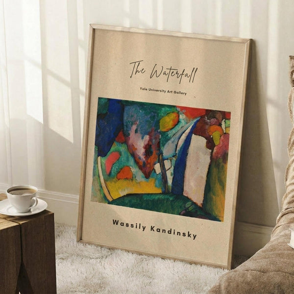 May include: A framed art print leaning against a white wall on a textured white rug. The poster features a colorful abstract painting with vibrant blues, greens, reds, yellows and white brushstrokes on a beige background with black text above and below the artwork. A white coffee cup sits on a wooden block nearby, and a beige knitted blanket is partially visible in the corner.