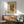 May include: Framed abstract art poster featuring vibrant brushstrokes in blues, greens, reds, and yellows on cream background, displayed above mid-century modern wooden sideboard with white ceramic lamp, next to sage green upholstered armchair with wooden frame in sunlit room with white walls