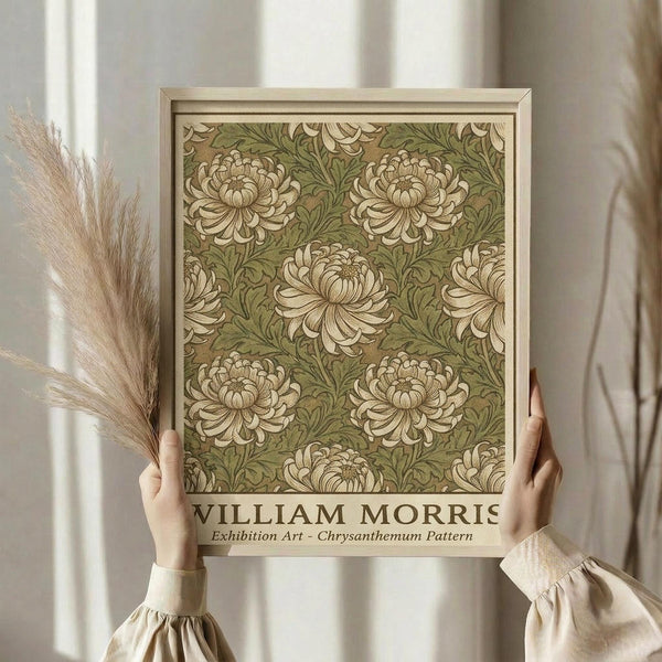 May include: Hands holding a framed botanical art print featuring cream chrysanthemum flowers with green foliage in William Morris's decorative pattern style. Light wooden frame against neutral curtain backdrop with dried pampas grass visible to the left. The artwork displays an elegant vintage floral wallpaper design with repeating blooms and ornate leaves in muted earth tones.
