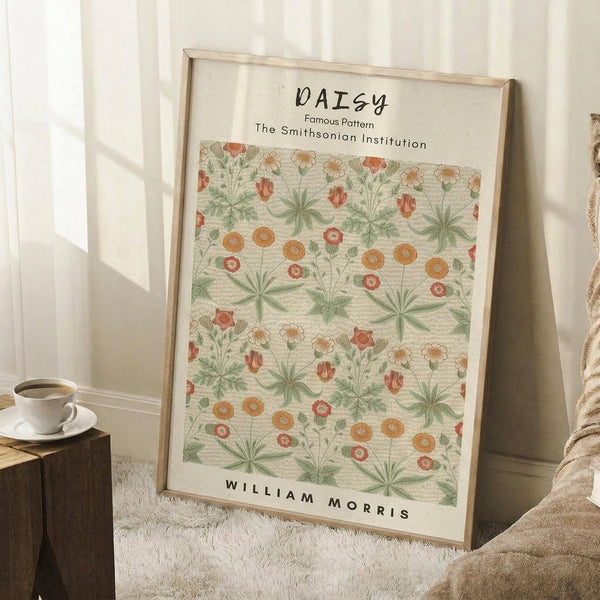 May include: A framed botanical print featuring William Morris's daisy pattern with green leaves, orange and coral flowers on cream background, displayed leaning against white wall corner on textured white rug. A coffee cup sits on wooden block nearby, with beige fabric visible at right edge.