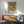 May include: Framed botanical art print featuring green and cream chrysanthemum floral pattern hanging on white wall above wooden sideboard. Mid-century modern living room with sage green upholstered armchair, round wood coffee table, and white ceramic table lamp. Natural sunlight creates geometric shadows across the minimalist Scandinavian-style interior.