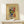 May include: Framed abstract art poster in light wood frame leaning against white wall on natural wood shelf. Artwork features vibrant colors including green, blue, red, yellow and orange in flowing brushstroke style. Small white wrapped gift box with evergreen sprig sits on shelf beside frame. Warm, minimal home decor setting with soft natural lighting.