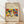 May include: Framed art print displaying a colorful abstract composition with bold red, yellow, blue, and green shapes featuring stylized human figures in an expressionist style. The print has cream-colored matting with handwritten-style text at top and bottom, housed in a light wood frame. The frame leans against a white wall on light wooden flooring with soft natural shadows cast across the scene.
