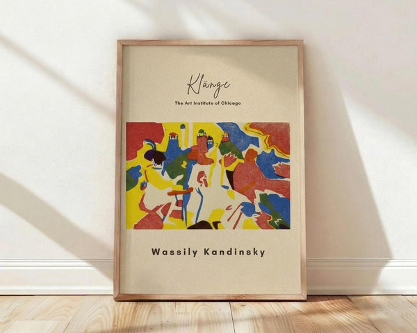 May include: Framed art print displaying a colorful abstract composition with bold red, yellow, blue, and green shapes featuring stylized human figures in an expressionist style. The print has cream-colored matting with handwritten-style text at top and bottom, housed in a light wood frame. The frame leans against a white wall on light wooden flooring with soft natural shadows cast across the scene.