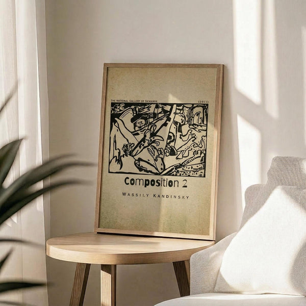 May include: Framed beige poster with black abstract line drawing displayed on round wooden side table next to white upholstered chair. Sunlight creates soft shadows across neutral-toned room with plant leaves visible in corner. Poster features bold, expressionist-style artwork with organic flowing forms and geometric elements in monochromatic design.