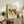 May include: Framed art print displaying abstract colorful figures in red, yellow, and blue tones, positioned on rustic wooden sideboard next to fern plant, books, and ceramic vase in warm-toned living room setting with cream upholstered chair visible.