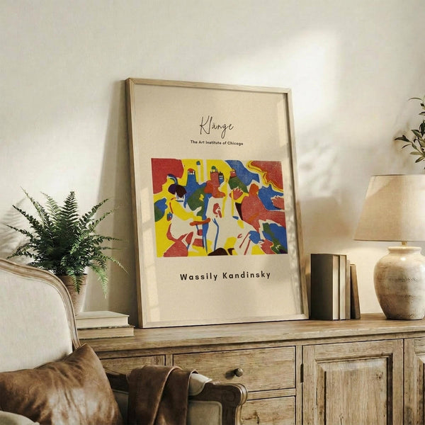 May include: Framed art print displaying abstract colorful figures in red, yellow, and blue tones, positioned on rustic wooden sideboard next to fern plant, books, and ceramic vase in warm-toned living room setting with cream upholstered chair visible.
