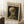 May include: Framed Van Gogh museum poster featuring skeleton with cigarette painting displayed on white wooden floor against white paneled wall, with coffee cup on wooden block nearby and beige textured blanket visible at edge. Poster has cream background with black text and shows skull smoking cigarette in Van Gogh's distinctive brushstroke style.