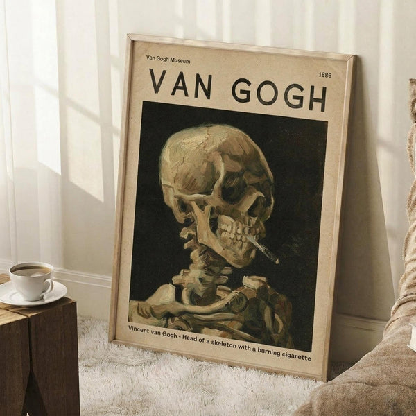 May include: Framed Van Gogh museum poster featuring skeleton with cigarette painting displayed on white wooden floor against white paneled wall, with coffee cup on wooden block nearby and beige textured blanket visible at edge. Poster has cream background with black text and shows skull smoking cigarette in Van Gogh's distinctive brushstroke style.