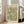 May include: Framed poster with vintage William Morris floral pattern in sage green and cream tones, featuring delicate botanical motif with flowers and leaves. Light wood frame leans against white wall next to beige upholstered armchair in bright, airy room with large windows and natural lighting on hardwood floors.