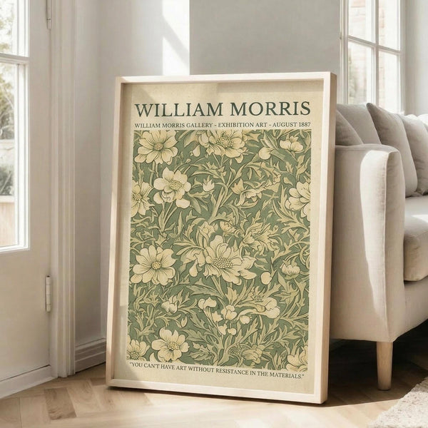 May include: Framed poster with vintage William Morris floral pattern in sage green and cream tones, featuring delicate botanical motif with flowers and leaves. Light wood frame leans against white wall next to beige upholstered armchair in bright, airy room with large windows and natural lighting on hardwood floors.