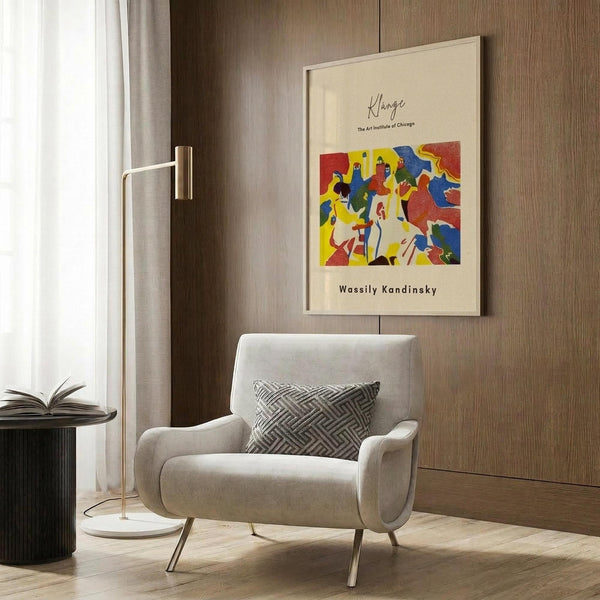May include: A framed vintage art exhibition poster featuring an abstract colorful painting with bold red, blue, yellow, and green shapes, displayed on a wood-paneled wall above a beige upholstered armchair with geometric patterned pillow. Modern floor lamp with brass finish stands nearby next to a dark round side table with open book. Neutral tones throughout the contemporary interior setting.