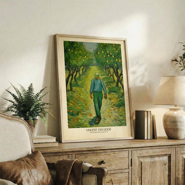 May include: Framed Van Gogh print showing a figure in blue clothing walking down a tree-lined path with green foliage and yellow fruit, displayed on rustic wooden sideboard alongside fern plant, ceramic vase, stacked books, and cream upholstered chair in neutral-toned living room setting.