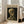 May include: Framed Van Gogh museum poster featuring skeleton with cigarette painting, displayed on light wooden floor against white wall next to beige upholstered chair. Print shows skull and ribcage in warm yellows and browns against dark background, with museum text and 1886 date. Natural sunlight streams through tall windows creating soft shadows on hardwood flooring.