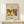 May include: Framed art print featuring Wassily Kandinsky's colorful abstract composition with bold shapes in red, yellow, blue, and green against cream background. Text reads "Klänge The Art Institute of Chicago" and "Wassily Kandinsky." Natural wood frame sits on light wooden surface next to small white gift box with greenery sprig, against neutral beige wall.