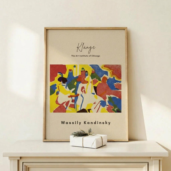 May include: Framed art print featuring Wassily Kandinsky's colorful abstract composition with bold shapes in red, yellow, blue, and green against cream background. Text reads "Klänge The Art Institute of Chicago" and "Wassily Kandinsky." Natural wood frame sits on light wooden surface next to small white gift box with greenery sprig, against neutral beige wall.