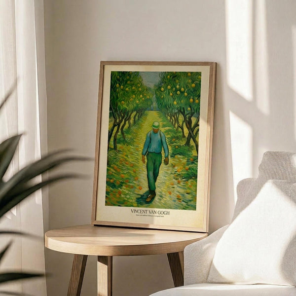 May include: Framed Van Gogh poster print displaying a man in blue jacket and green pants walking down a tree-lined path with yellow fruit trees, shown on round wooden side table next to beige upholstered chair in sunlit room with white walls and plant shadows
