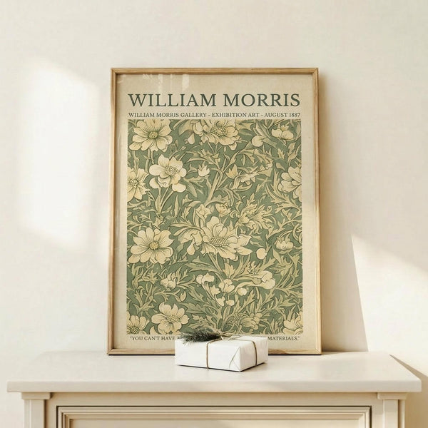 May include: Framed vintage botanical poster featuring William Morris floral pattern in sage green and cream tones, displayed on light wood console table beside small white gift box with dried sprig, against neutral wall background.