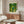 May include: Vincent Van Gogh poster print displayed above wooden credenza in modern living room. Artwork shows figure in blue shirt and green pants walking through orchard with yellow fruit trees. Warm wood furniture, white curved chair, glass coffee table, and soft natural lighting create contemporary interior setting.