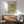 May include: Framed William Morris botanical poster with green floral pattern hanging above wooden credenza. Mid-century modern living room setting with sage green upholstered armchair, white ceramic table lamp, and round wood coffee table. Natural sunlight creates geometric shadows on white wall. Vintage-inspired exhibition art print in thin brown frame showcases intricate leaf and flower motifs in muted green tones.
